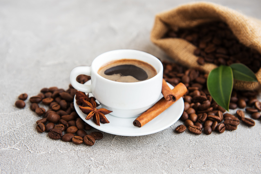 Tasse Kaffee mit Zimtstange, Sternanis und Kaffeebohnen neben einem Jutesack auf hellem Untergrund