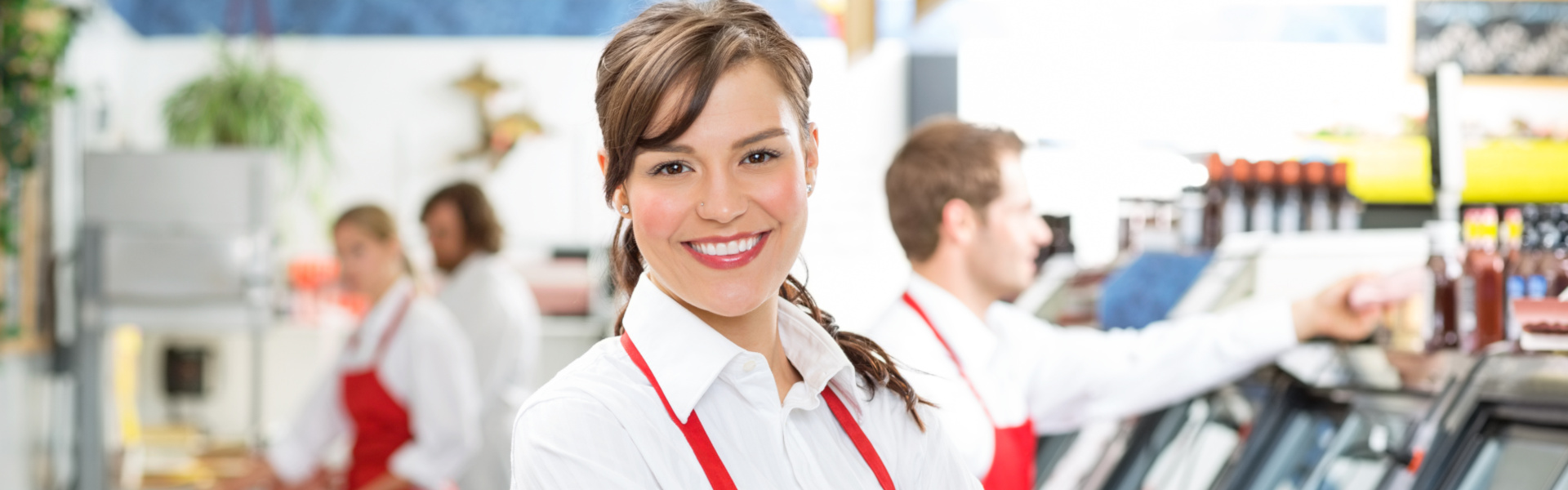 junge Mitarbeiterin in roter Schürze steht lächelnd mit verschränkten Armen in einem Lebensmittelgeschäft